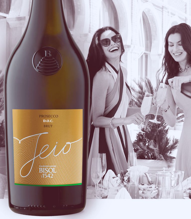 A bottle of Jeio Cuvée Brut Prosecco by Bisol stands in the foreground to the left. The background is a black and white scene of two women at a resort, wearing flowy resort dresses, pouring Jeio Cuvée Brut into champagne glasses and having a laugh.