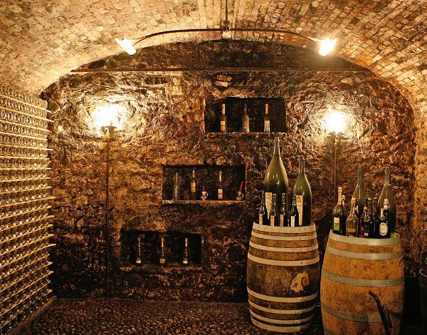 The underground cellar of Cantine Bisol: a small, low-lit stone room with wine bottles stacked on their side to the left, previous vintage wines standing atop 2 barrels to the right, as well as in 3 upward diagonal hollows in the back wall.