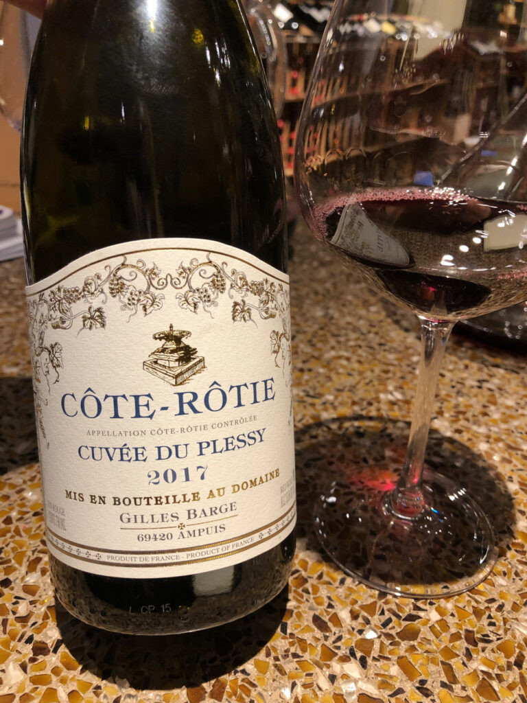 The bottom two thirds of a bottle of Côte-Rôtie Cuvee du Plessy is photographed to left of a wine glass with a tasting of the wine. Both sit atop a mustard and brown coloured terrazzo countertop.