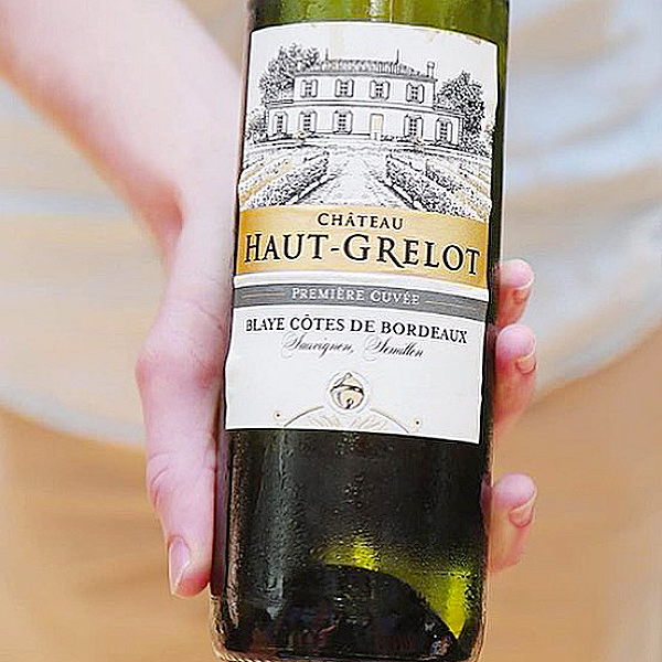 An image of a male caucasian hand presenting a bottle that reads Chateau Haut-Grelot Première Cuvée, Blaye Côtes de Bordeaux, and two unidentifiable words printed in calligraphy.