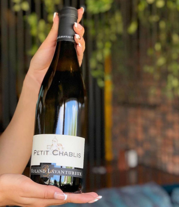 An image of caucasian female hands with french tip manicure brandishing a bottle of Petit Chablis by Roland Lavantureux. A garden lounge is out of focus in the background. 